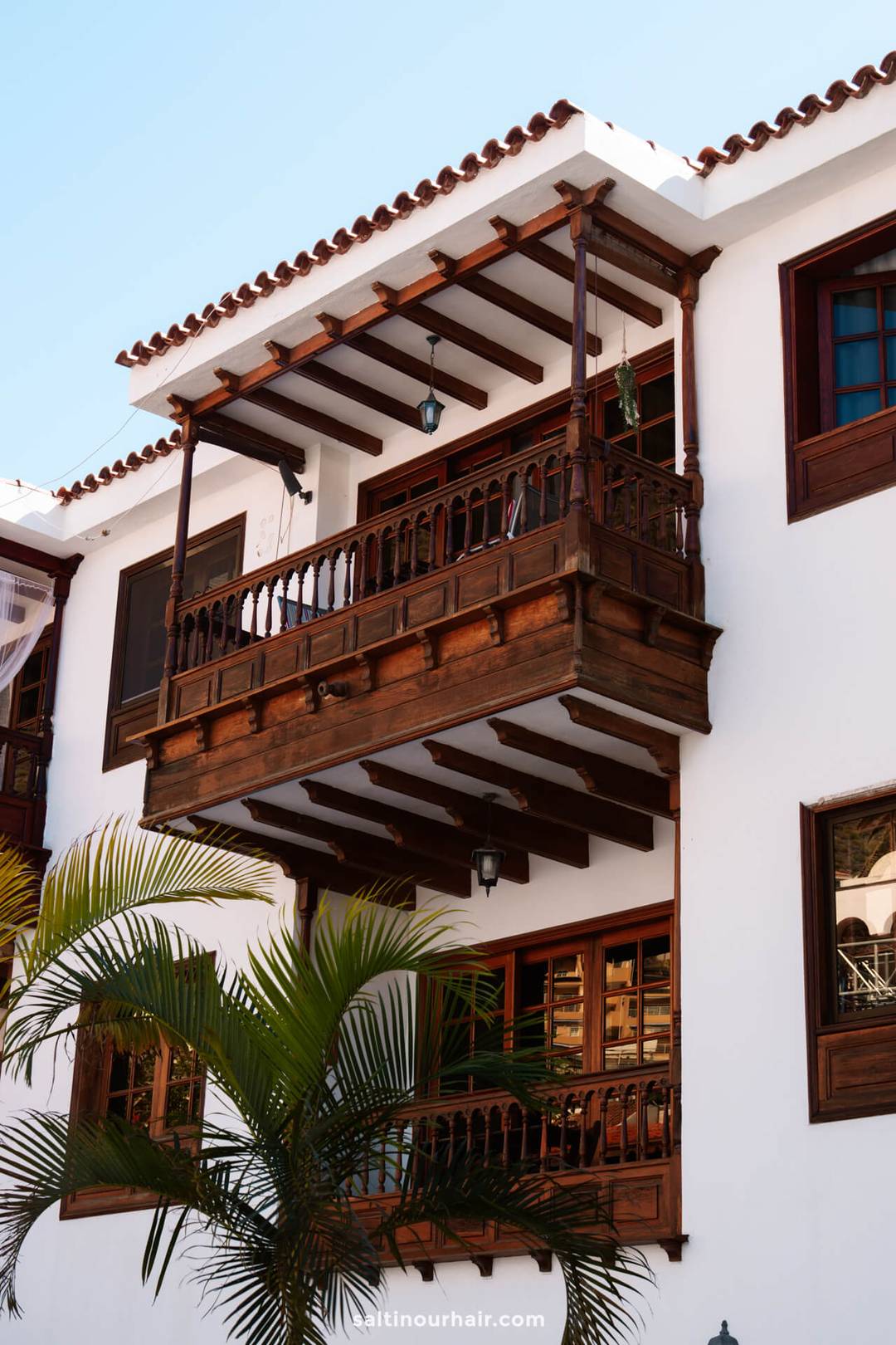 A traditional white building with wooden balconies and windows, featuring a tiled roof and a potted palm in the foreground, captures the charm of Los Gigantes village Tenerife.