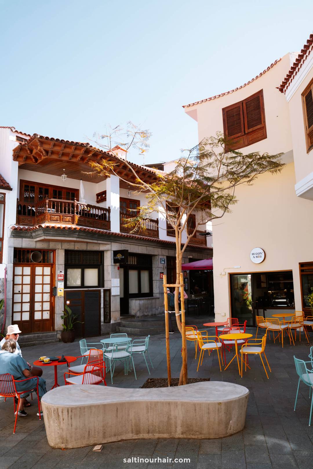 Outdoor caf&eacute; scene with colorful chairs and tables, a curved concrete bench, and a tree in a town square bordered by white and yellow buildings with balconies, capturing the charming ambiance of Los Gigantes village Tenerife.