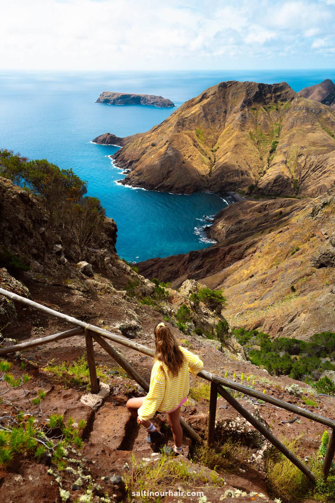A woman in a yellow striped shirt stands on a wooden stairway along the PSPR1 hike, overlooking Porto Santo Madeira&rsquo;s rugged coastline with cliffs and blue ocean below.