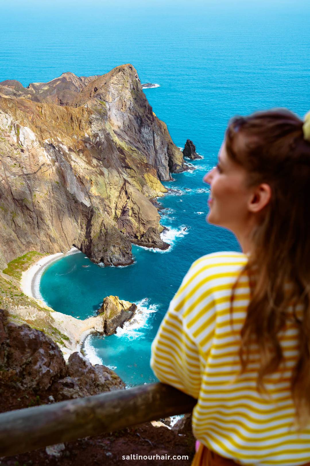 A woman in a yellow striped shirt looks out over Porto Santo&rsquo;s rocky coastline with turquoise water and steep cliffs, capturing the beauty of one of the top things to do in Porto Santo.