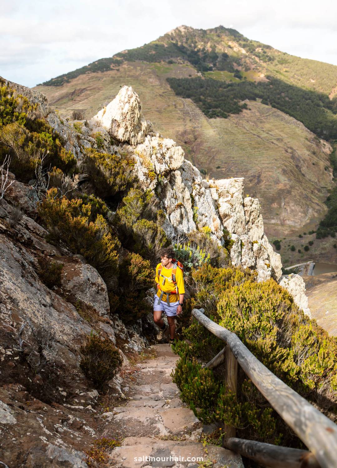 A man wearing a yellow backpack hikes up a narrow rocky trail with a wooden railing, surrounded by mountains and shrubs&mdash;a perfect scene among the many things to do in Porto Santo.