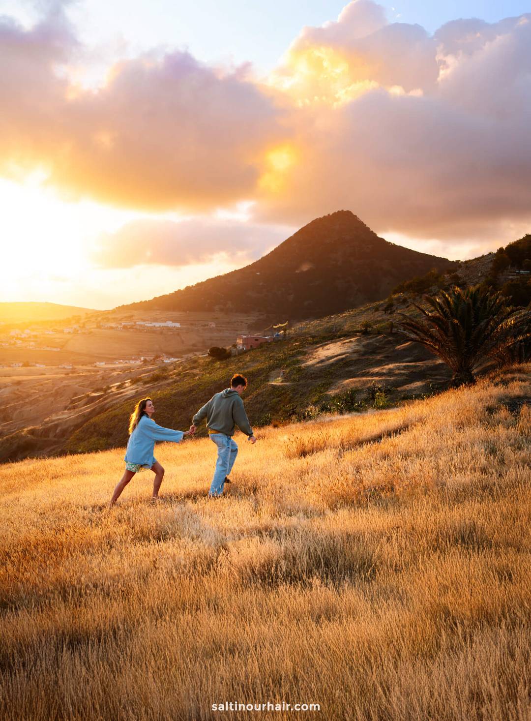 Twee mensen lopen hand in hand door een gouden veld bij zonsondergang, met een berg en indrukwekkende wolken op de achtergrond &mdash; een moment van avontuur zonder zorgen over de wandelkosten op Madeira.