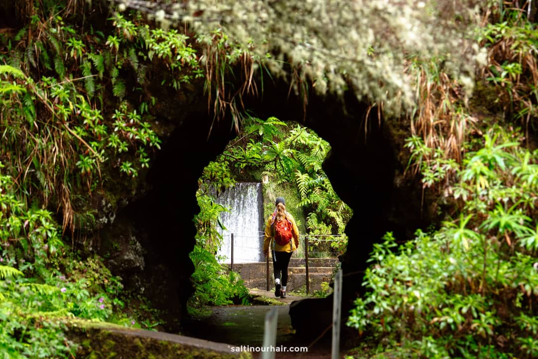 Een persoon met een rode rugzak loopt door een tunnel omringd door weelderige groene vegetatie, met een waterval op de achtergrond &mdash; een onvergetelijke sc&egrave;ne op de beroemde paden van het eiland waar wandelkosten van toepassing kunnen zijn.