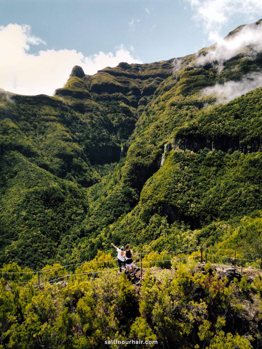 Twee mensen staan op een bergpad omringd door weelderig groene heuvels en valleien onder een halfbewolkte hemel, terwijl ze een van de mooiste wandelingen op Madeira maken - de beroemde PR17-route.