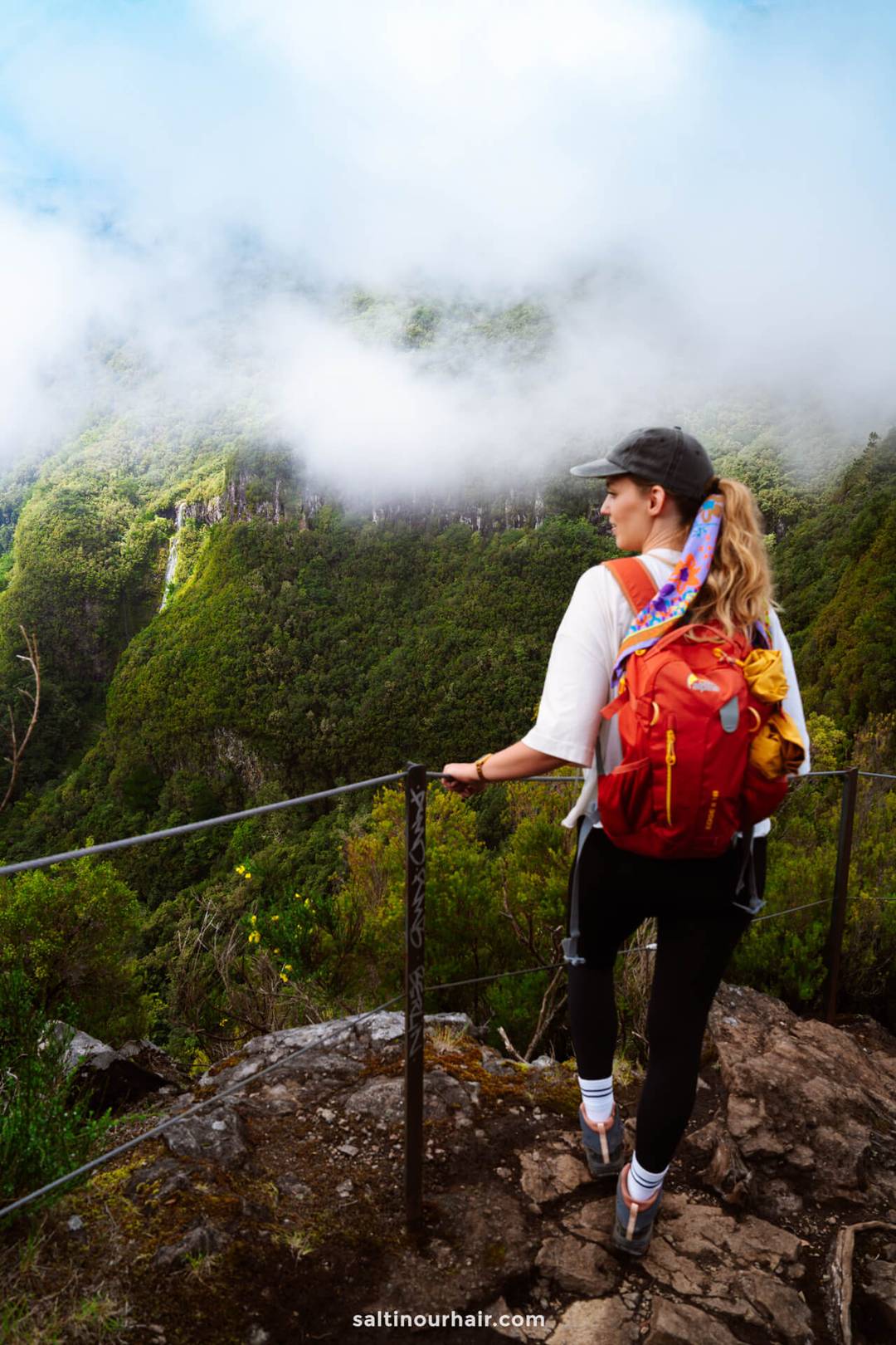 Een vrouw met een rode rugzak staat op een rotsachtig pad met uitzicht op een weelderige, groene vallei die gedeeltelijk bedekt is door wolken tijdens de pr17 hike - een van de mooiste hikes van madeira