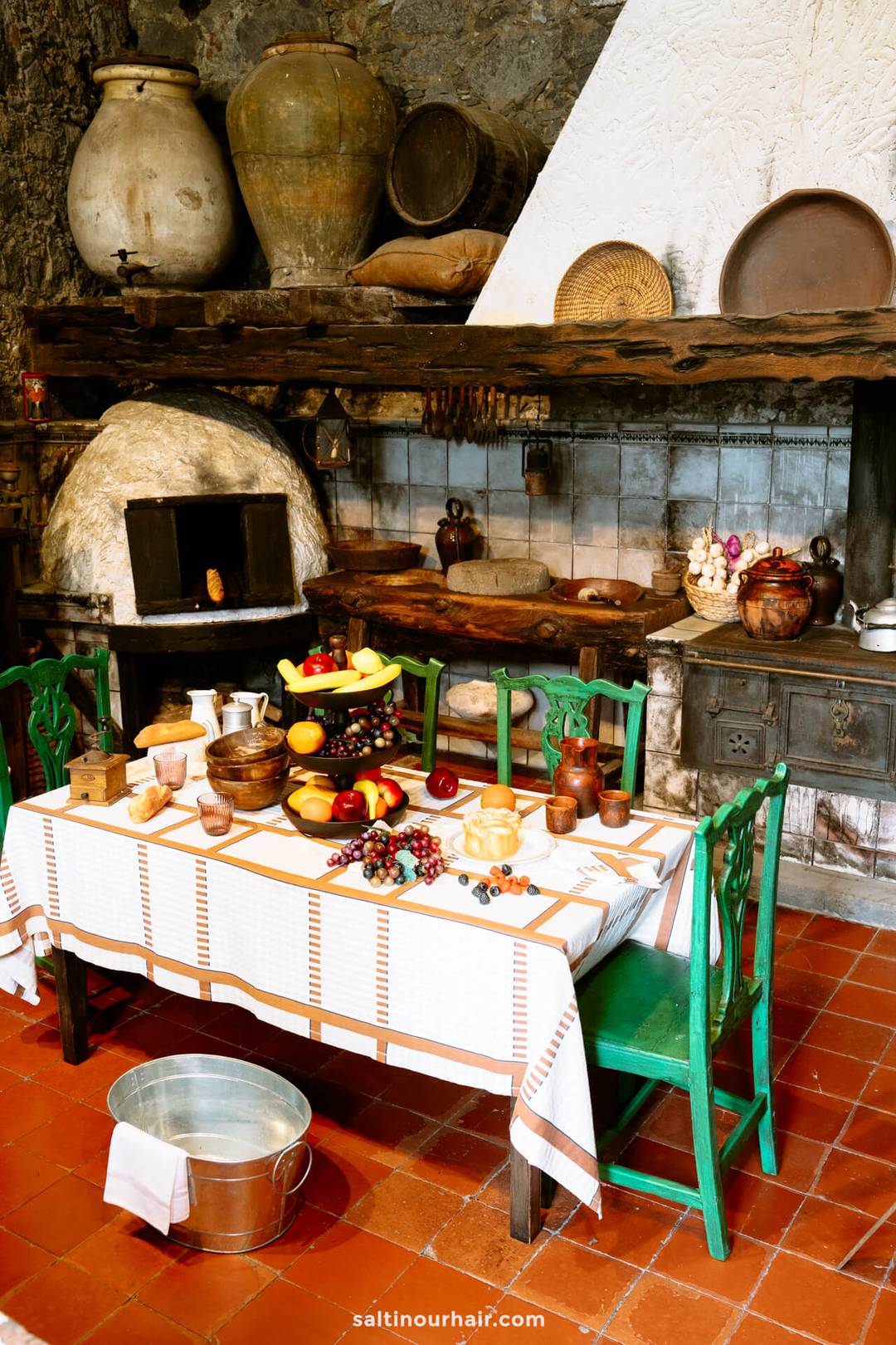 A rustic kitchen in Puerto de la Cruz with a tiled floor, wooden shelves, clay pots, and an oven. A table set with fruit, bread, and dishes stands surrounded by green chairs.