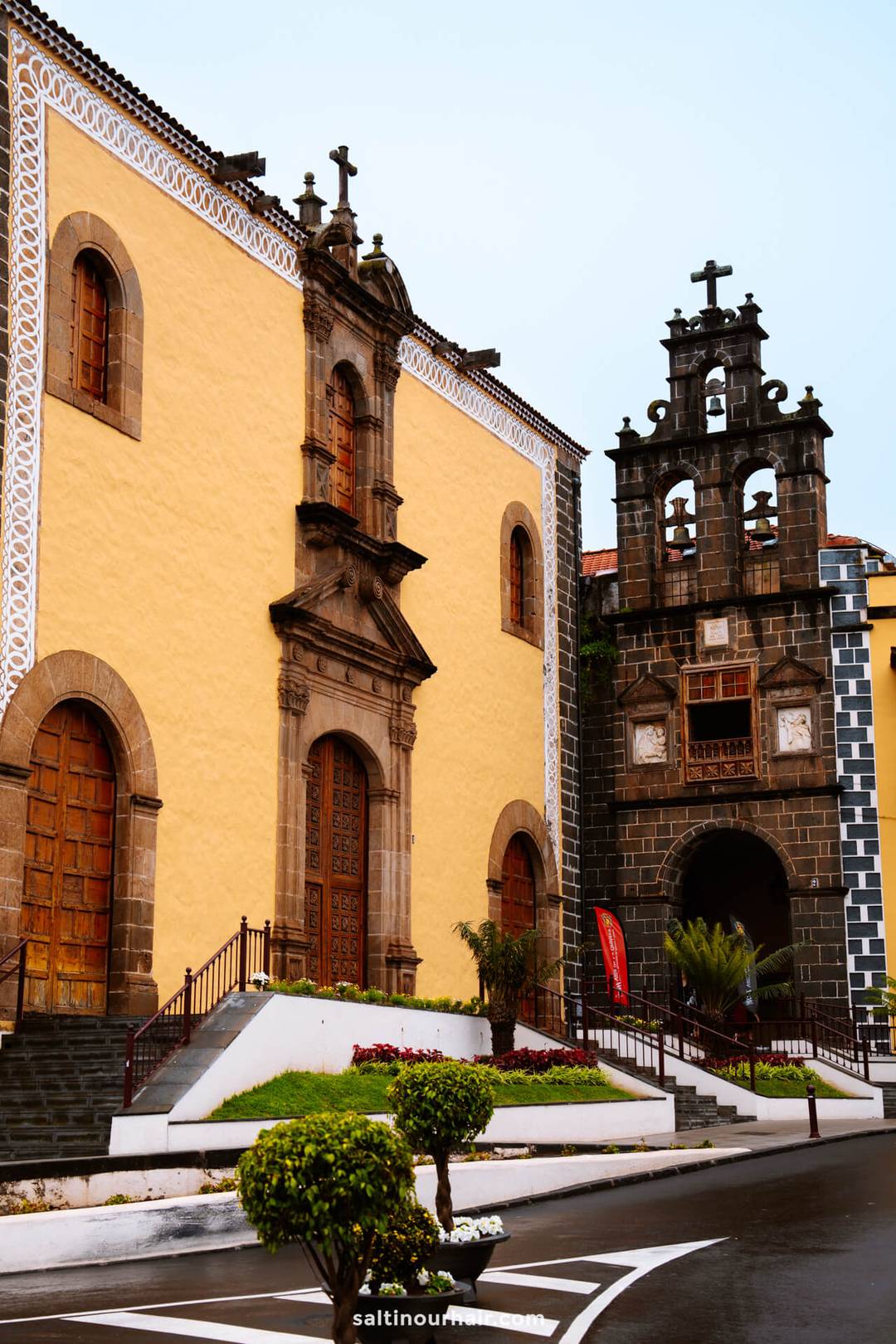 A historic church in la orotava with a yellow facade, arched wooden doors, and an adjacent bell tower adorned with multiple crosses and clocks, set amid landscaped urban surroundings.