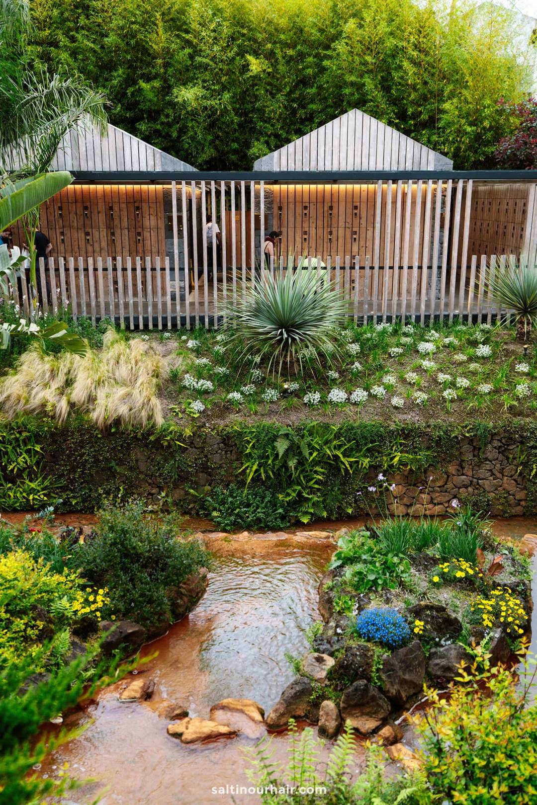 Modern wooden building with vertical slats nestled behind a lush garden, featuring green plants, flowers, and a small stream&mdash;hot springs in S&atilde;o Miguel