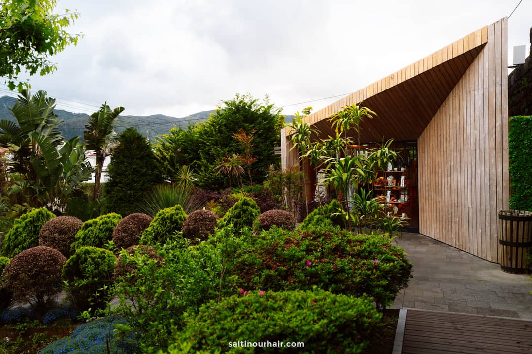 A modern wooden building with large windows is surrounded by manicured bushes, trees, and lush green landscaping, reminiscent of the relaxing retreats near the hot springs in S&atilde;o Miguel