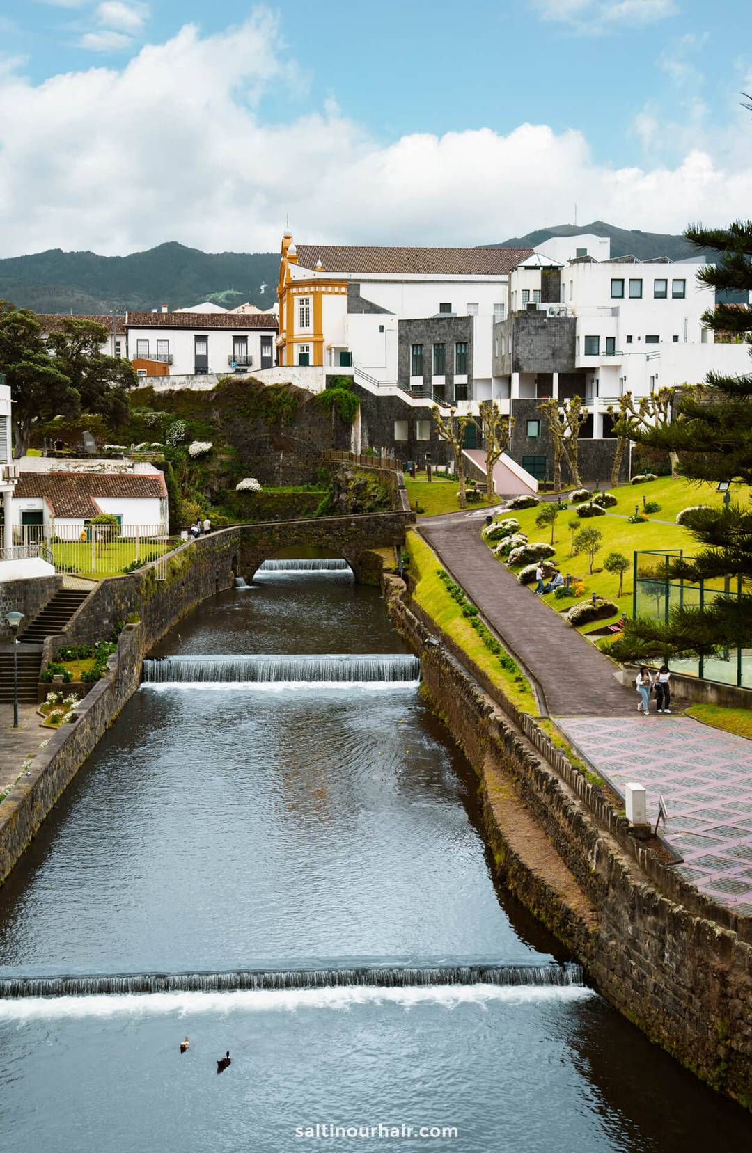 A narrow river with small waterfalls flows through a landscaped town called Ribeira Grande with white buildings, a yellow church, and green hills in the background&mdash;an inviting stop on any S&atilde;o Miguel itinerary.