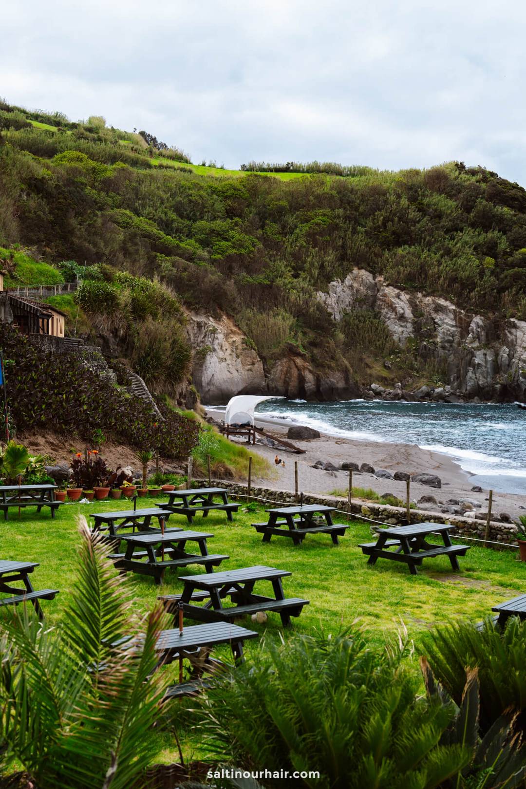 Picnic tables on a grassy area overlook a rocky beach and ocean, with green hills and cliffs in the background under a cloudy sky&mdash;an ideal stop on any S&atilde;o Miguel itinerary.