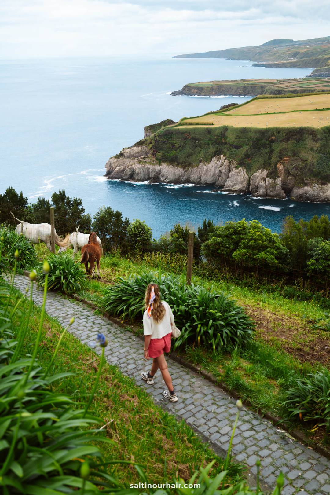 A person walks on a stone path through lush greenery near a coastal cliff, with two horses grazing nearby and the ocean in the background&mdash;a perfect moment for any S&atilde;o Miguel itinerary.
