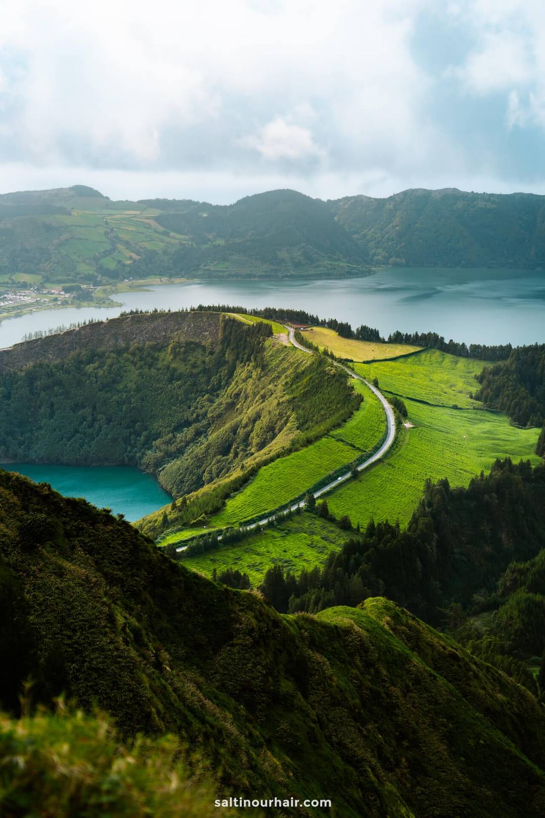 A winding road cuts through green hills overlooking a lake, perfect for any S&atilde;o Miguel itinerary, with mountains and a cloudy sky in the background.