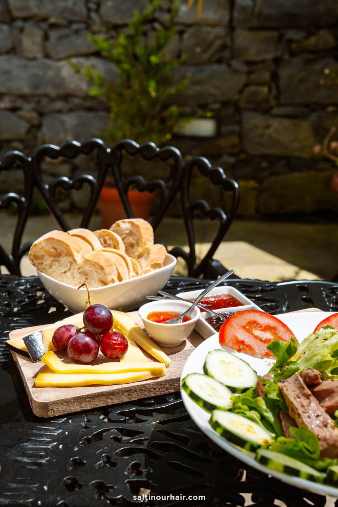 A table set outdoors with sliced bread, grapes, cheese, spreads, and a salad with cucumber, tomato, and lettuce&mdash;perfect for a relaxing pause during your S&atilde;o Miguel itinerary. Black metal chair and stone wall in the background.