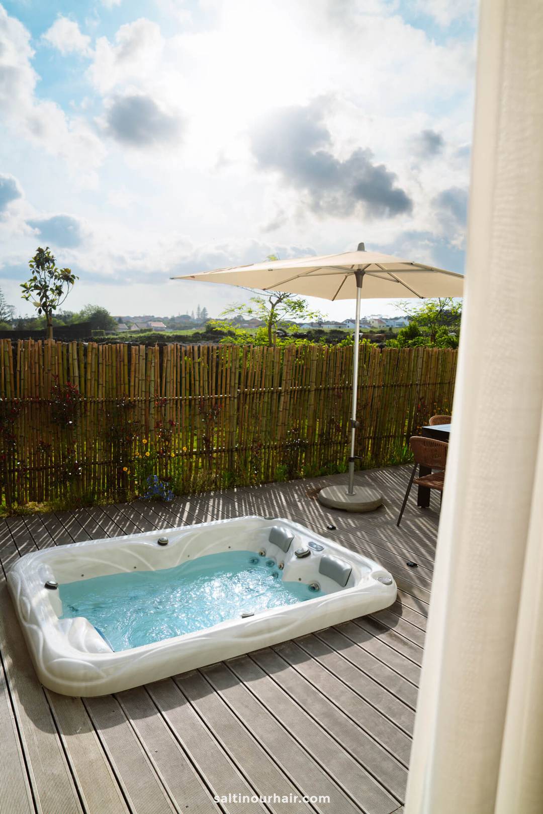 A white outdoor hot tub sits on a wooden deck with bubbling water, next to a sun umbrella, bordered by a bamboo fence. the perfect hotel in ponta delgada sao miguel azores