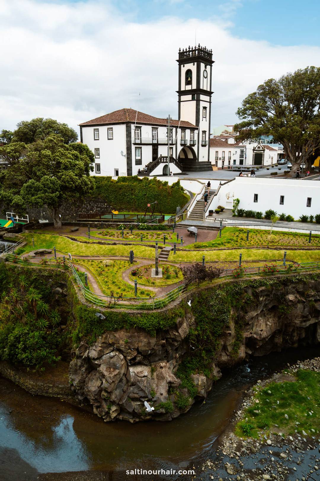 White building with a clock tower in Ribeira Grande beside a lush garden and footbridge, overlooking a rocky stream under a partly cloudy sky&mdash;an ideal stop on any S&atilde;o Miguel itinerary.