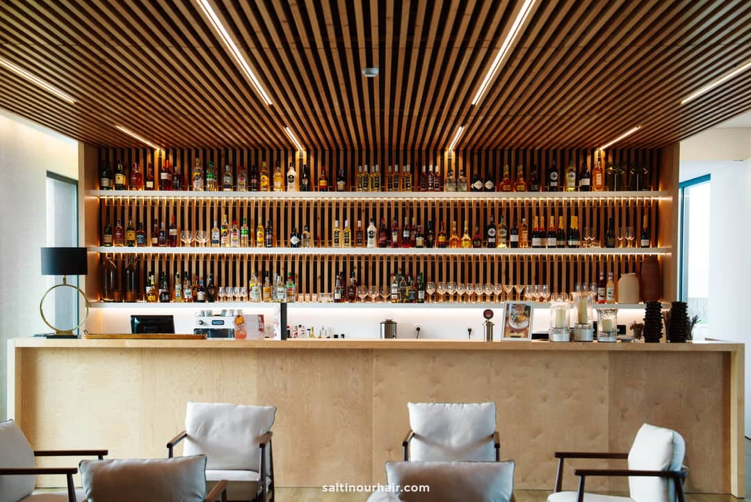 A modern bar with wooden accents at Lusitano Garden Villas, perfect for relaxing during your S&atilde;o Miguel itinerary, features a well-stocked back wall of liquor bottles, a counter with drinkware, and white cushioned chairs in the foreground.