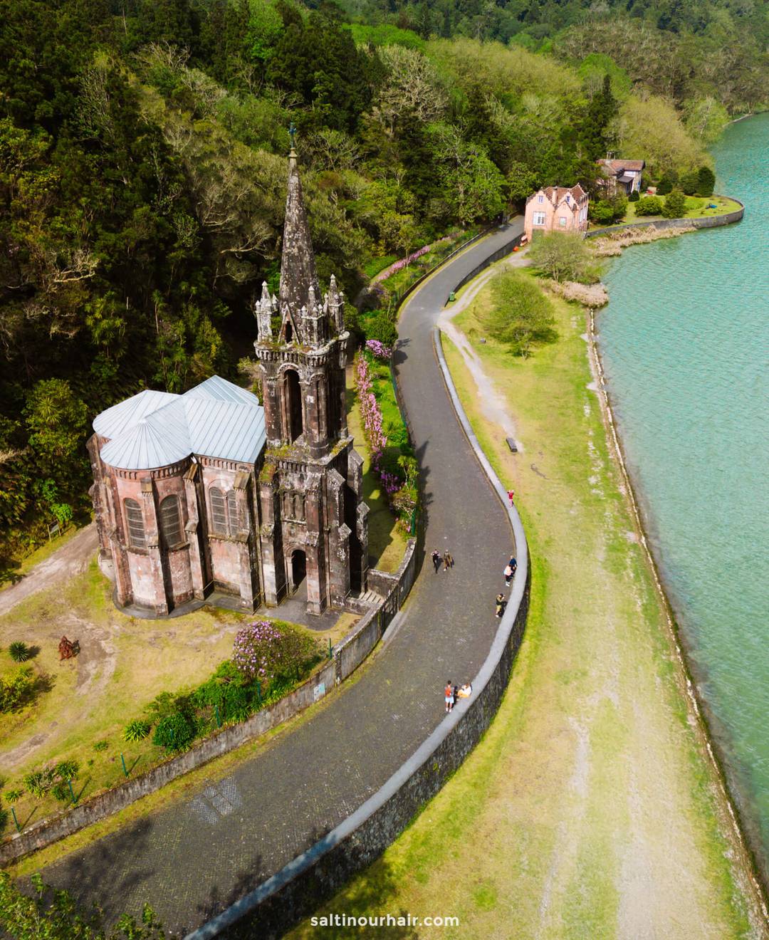 Aerial view of a historic stone church called Capela de Nossa Senhora das Vit&oacute;rias with a tall spire beside a winding road next to a turquoise lake, surrounded by greenery&mdash;a must-see highlight on any S&atilde;o Miguel itinerary.
