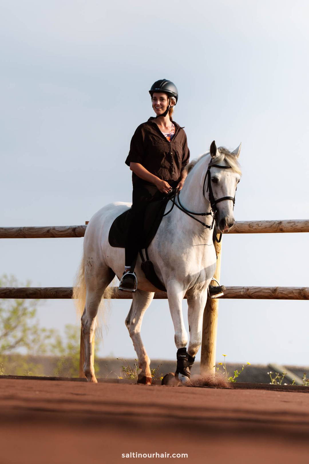 A woman wearing a helmet rides a white horse along a fenced dirt path outdoors, one of the many adventurous things to do in Ponta Delgada sao miguel azores