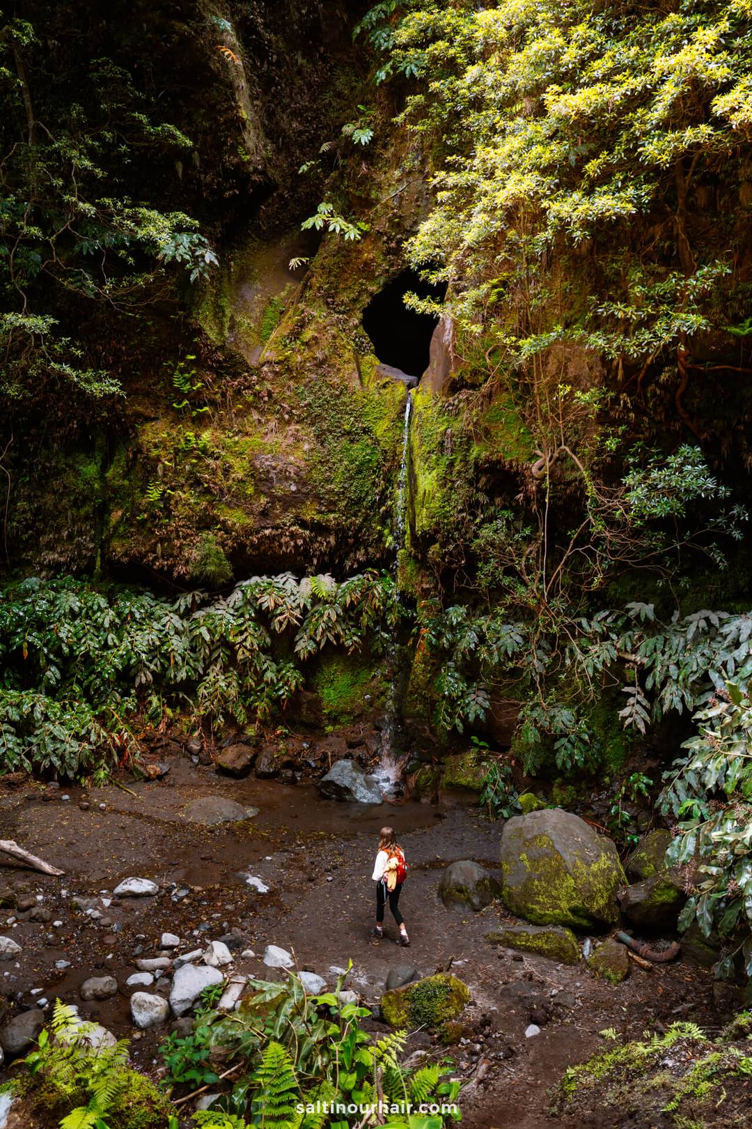 A woman with a backpack stands on rocky ground near  Janela do Inferno Waterfall flowing from a circular hole in a moss-covered cliff, surrounded by dense green foliage&mdash;an adventurous moment among the many things to do in S&atilde;o Miguel, Azores.