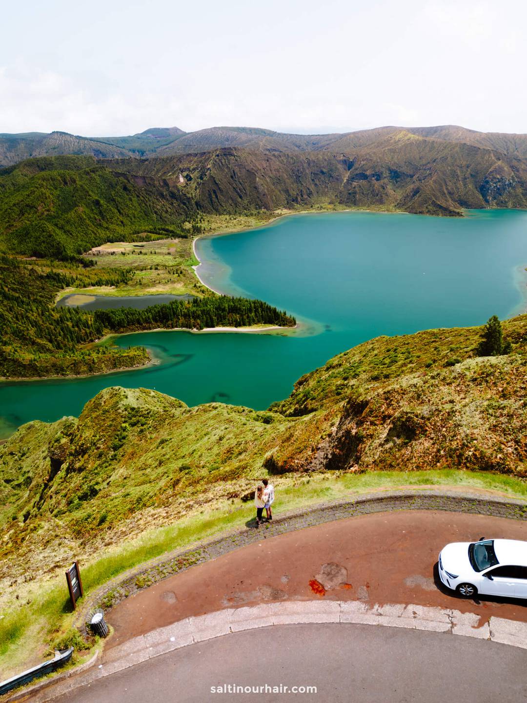 A couple stands on a curved road overlooking a turquoise lake and green mountains called Lagoa do Fogo, with a white car parked nearby&mdash;capturing one of the many unforgettable things to do in S&atilde;o Miguel, Azores