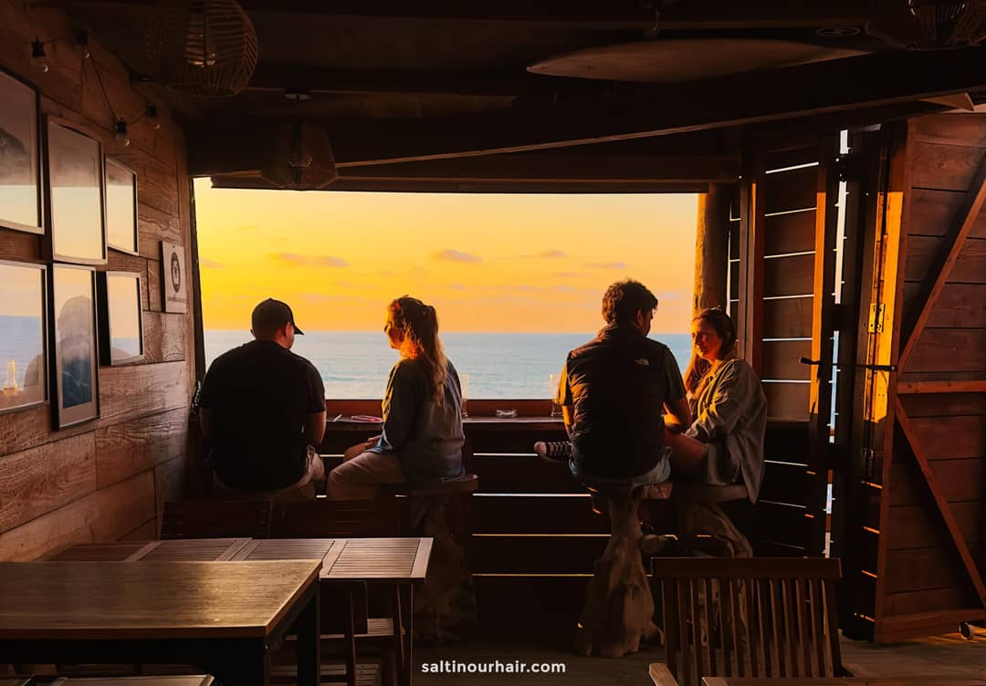 Four people sit at a wooden counter inside a rustic cabin, looking out at the ocean and sunset through an open window&mdash;a perfect way to unwind after exploring things to do in S&atilde;o Miguel, Azores.