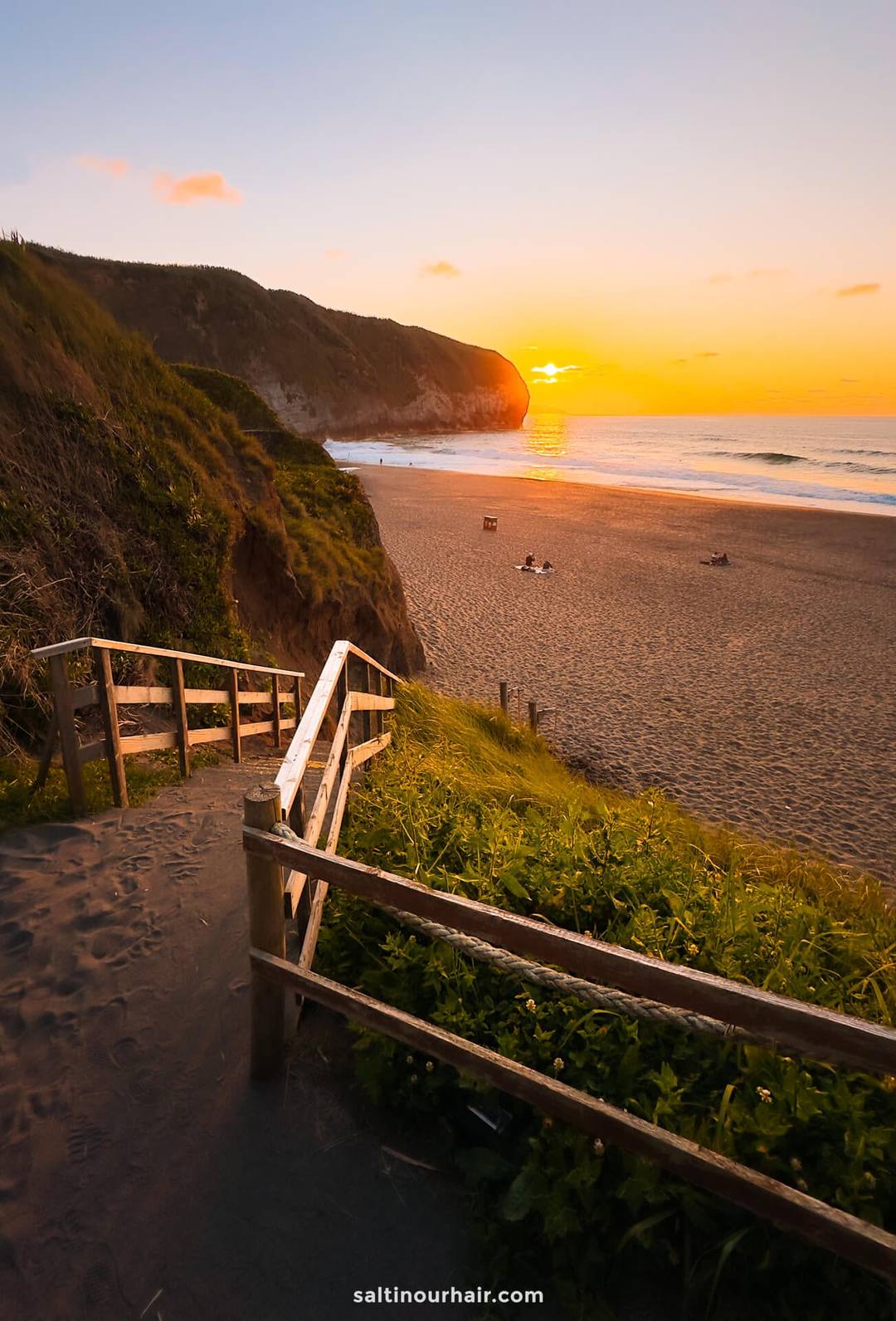 A sandy pathway with wooden railings leads down to Santa B&aacute;rbara Beach at sunset, with cliffs on one side and a few people sitting on the shore&mdash;a perfect spot for those seeking unique things to do in S&atilde;o Miguel, Azores.