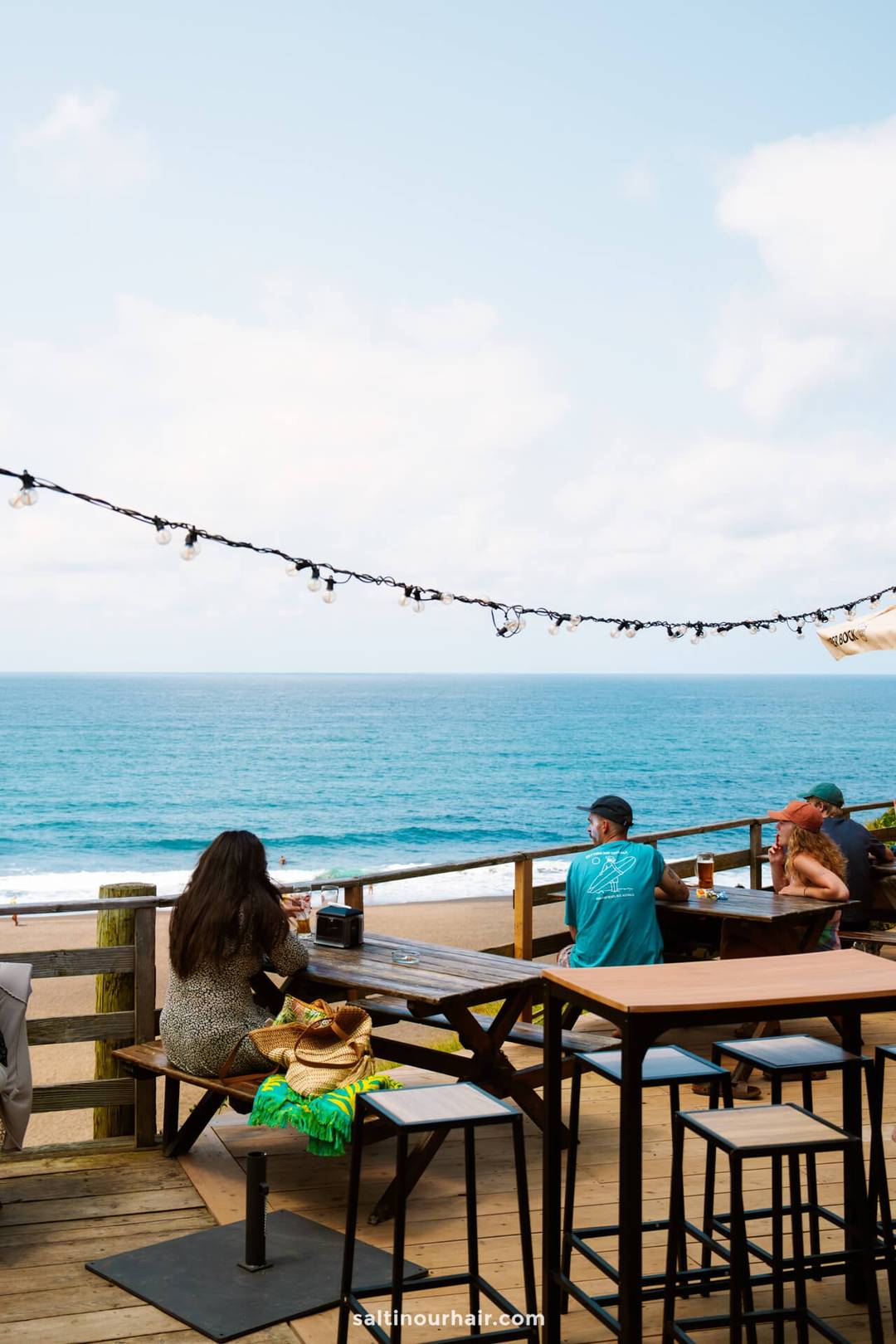 People sit at wooden tables on a deck overlooking the ocean under a partly cloudy sky with string lights above&mdash;a perfect spot to unwind and enjoy one of the best things to do in S&atilde;o Miguel, Azores.