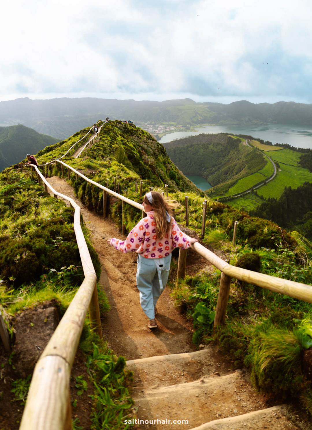 A woman walks down a dirt path at Sete Cidades Miradouro da Grota do Inferno with wooden railings along a scenic, green mountain ridge overlooking a lake and valley below&mdash;a perfect example of the breathtaking things to do in S&atilde;o Miguel, Azores.