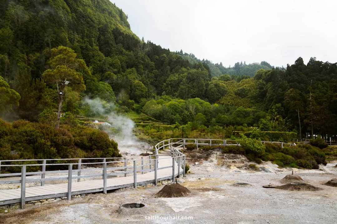 A wooden boardwalk winds through a geothermal area with steam rising from the ground, surrounded by dense green forest and hills&mdash;one of the must-see things to do in S&atilde;o Miguel, Azores.
