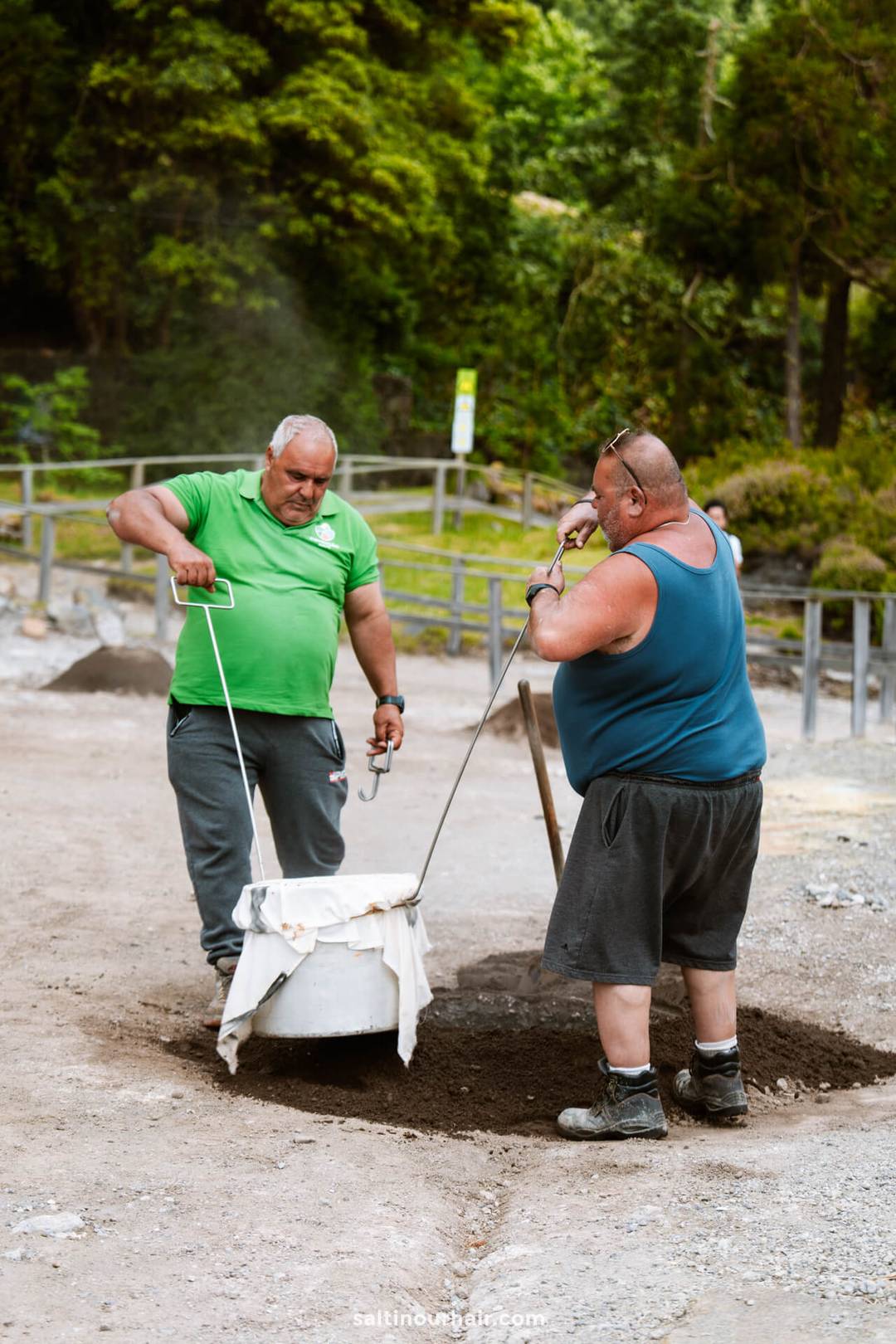 Two men stand on dirt, lifting a large pot out of the ground using metal rods&mdash;a traditional scene among the trees and wooden fence. Discovering this is one of the unique things to do in S&atilde;o Miguel, Azores.