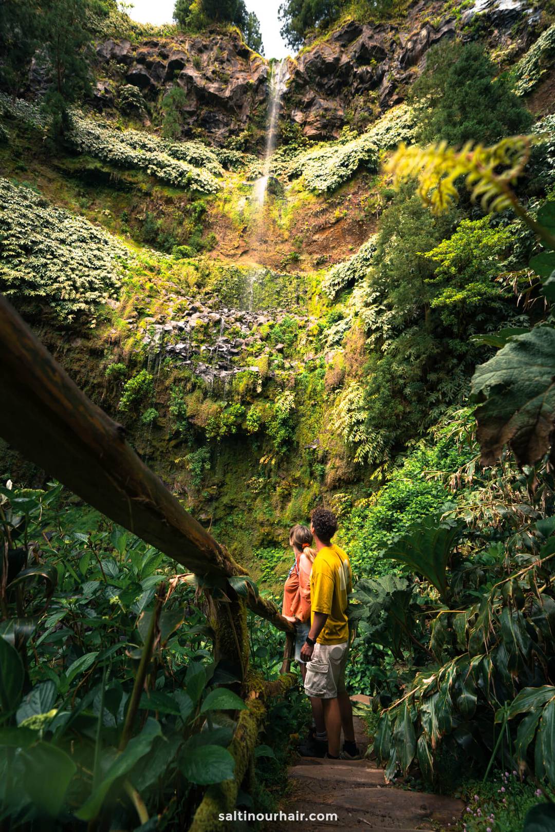 A young couple stand on a wooden path in Parque da Gren&aacute; surrounded by dense green foliage, gazing up at a tall, thin waterfall cascading down a lush, rocky cliff&mdash;one of the unforgettable things to do in S&atilde;o Miguel, Azores.