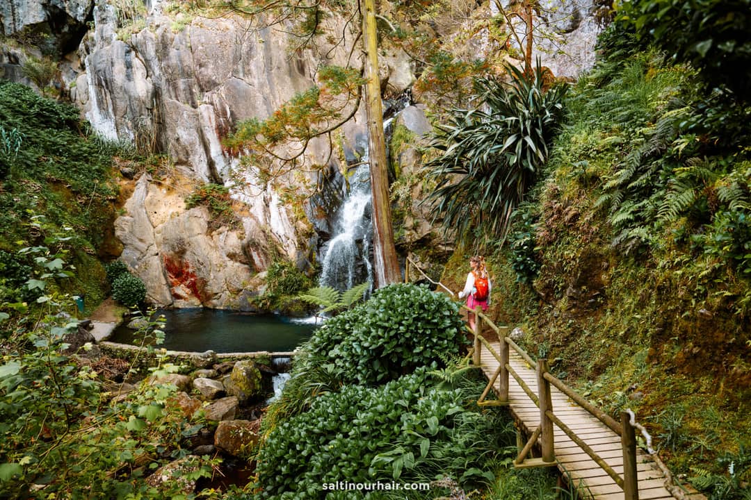 A person stands on a wooden bridge surrounded by dense greenery, facing a small waterfall flowing into a pool at the base of rocky cliffs&mdash; Cascata do Limbo is the perfect spot for those seeking adventures things to do in S&atilde;o Miguel, Azores.