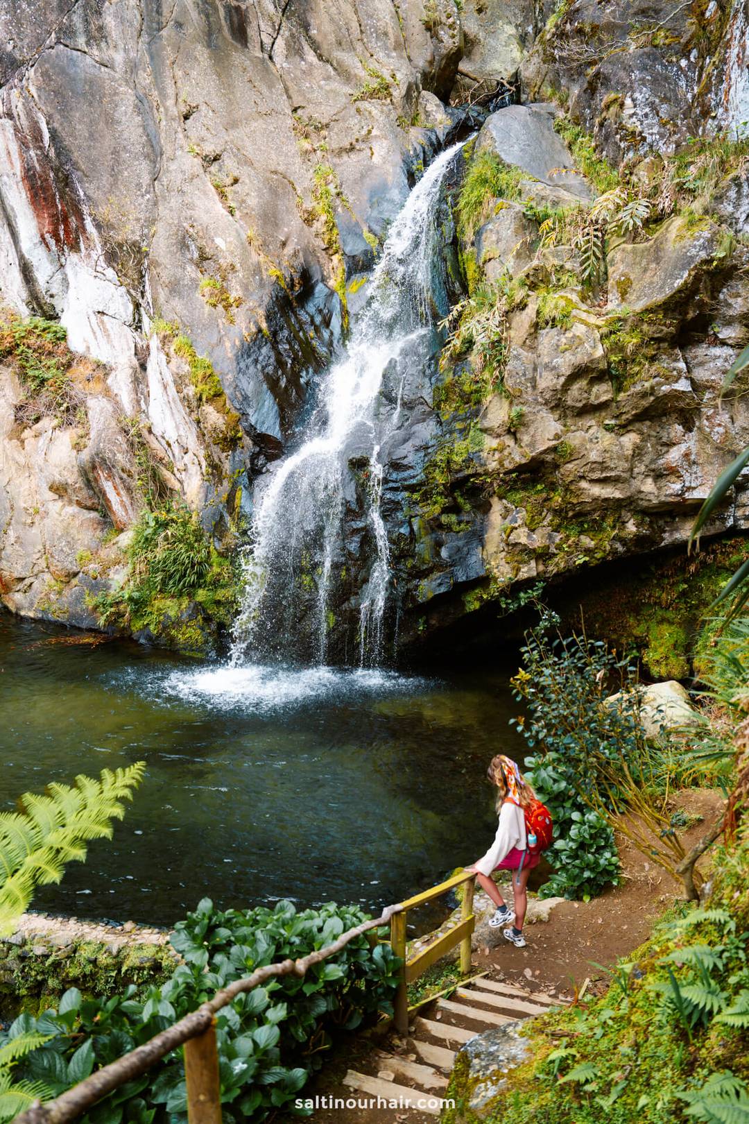 A woman with a red backpack stands on a wooden path next to a small waterfall called Cascata do Limbo cascading into a rocky pool surrounded by greenery, capturing one of the must-see things to do in S&atilde;o Miguel.