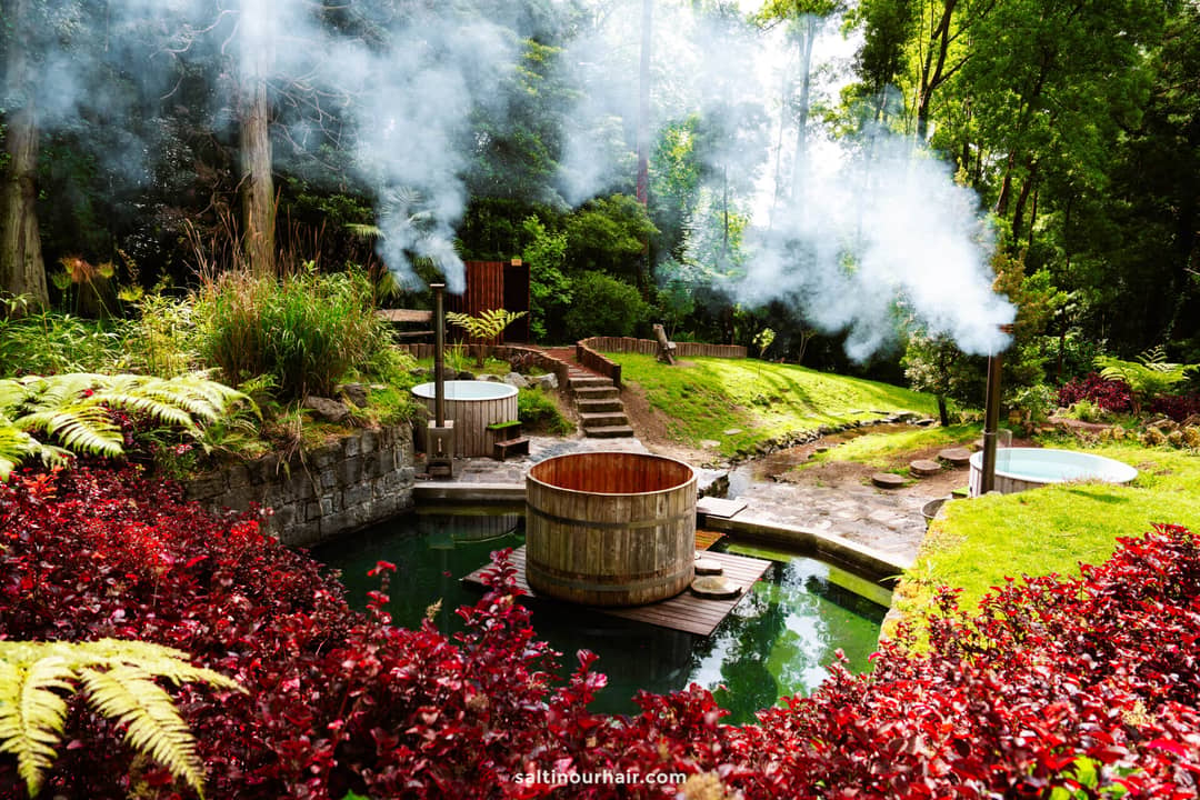 Three wooden hot tubs with steam rising, surrounded by lush greenery and red foliage in a forest setting&mdash;one of the most relaxing things to do in S&atilde;o Miguel.