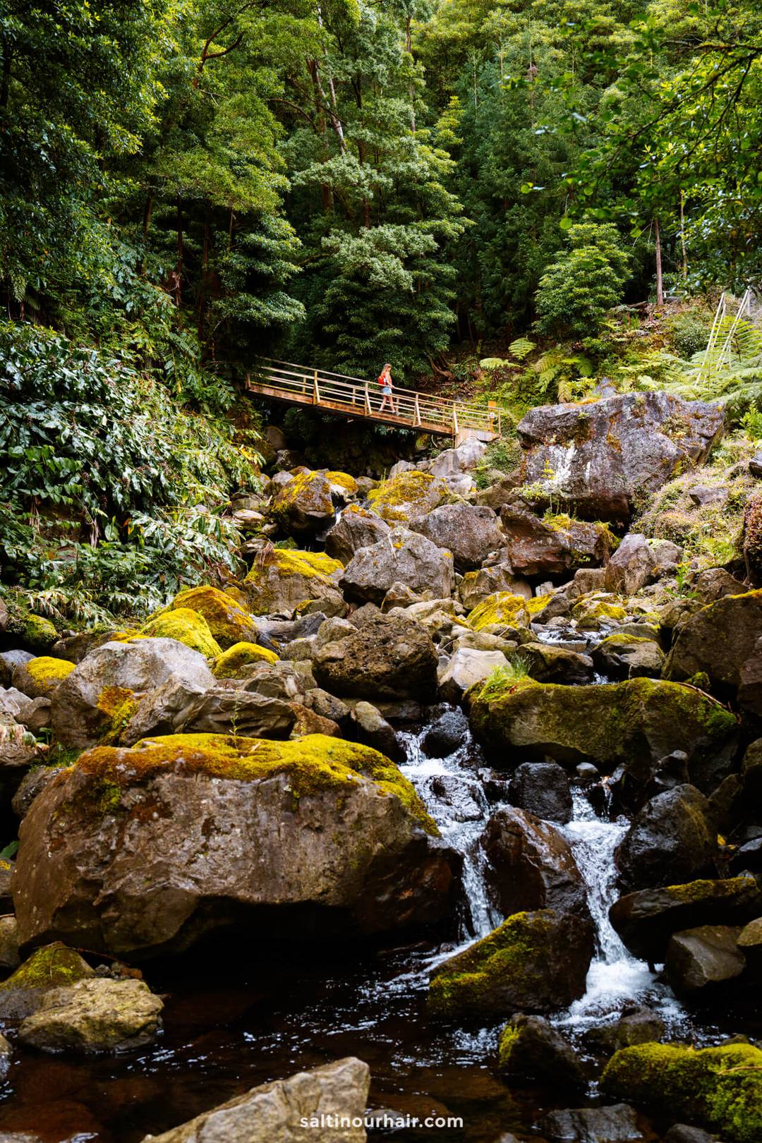 A woman walks over a small wooden bridge that crosses over a rocky creek with moss-covered boulders, surrounded by dense, green forest in Parque da Gren&aacute;&mdash;one of the enchanting things to do in S&atilde;o Miguel, Azores.