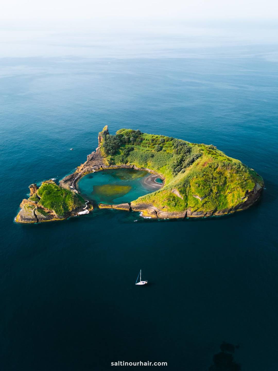 A small, green, rocky island with a central lagoon sits in calm blue ocean waters; a single sailboat floats nearby&mdash;evoking the serene beauty and tranquil escapes found among the best things to do in S&atilde;o Miguel, Azores.