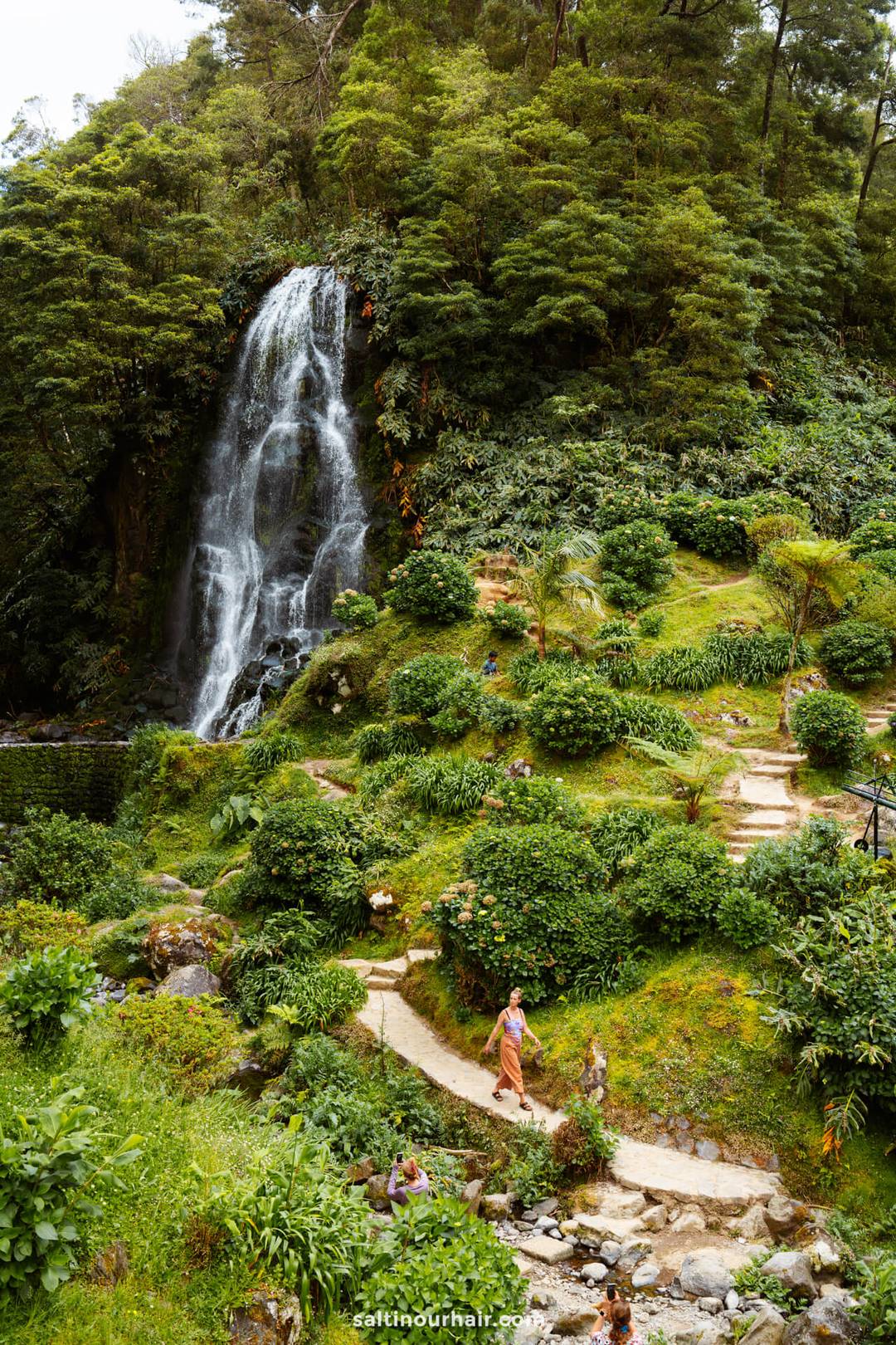 A tall waterfall flows down a cliff surrounded by dense green foliage at Parque Natural da Ribeira dos Caldeir&otilde;es, with people walking along a winding stone path below&mdash;a must-see among the top things to do in S&atilde;o Miguel, Azores.