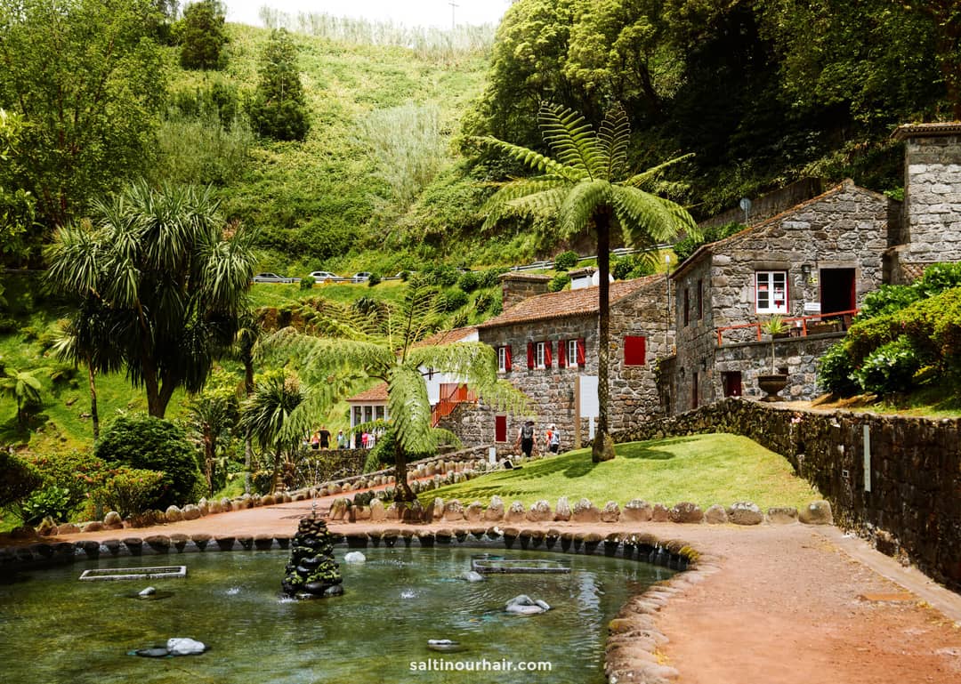 A stone house with red shutters sits beside a pond and lush greenery at Parque Natural da Ribeira dos Caldeir&otilde;es, where people walk along a path in this hilly, forested area&mdash;one of the scenic things to do in S&atilde;o Miguel, Azores.