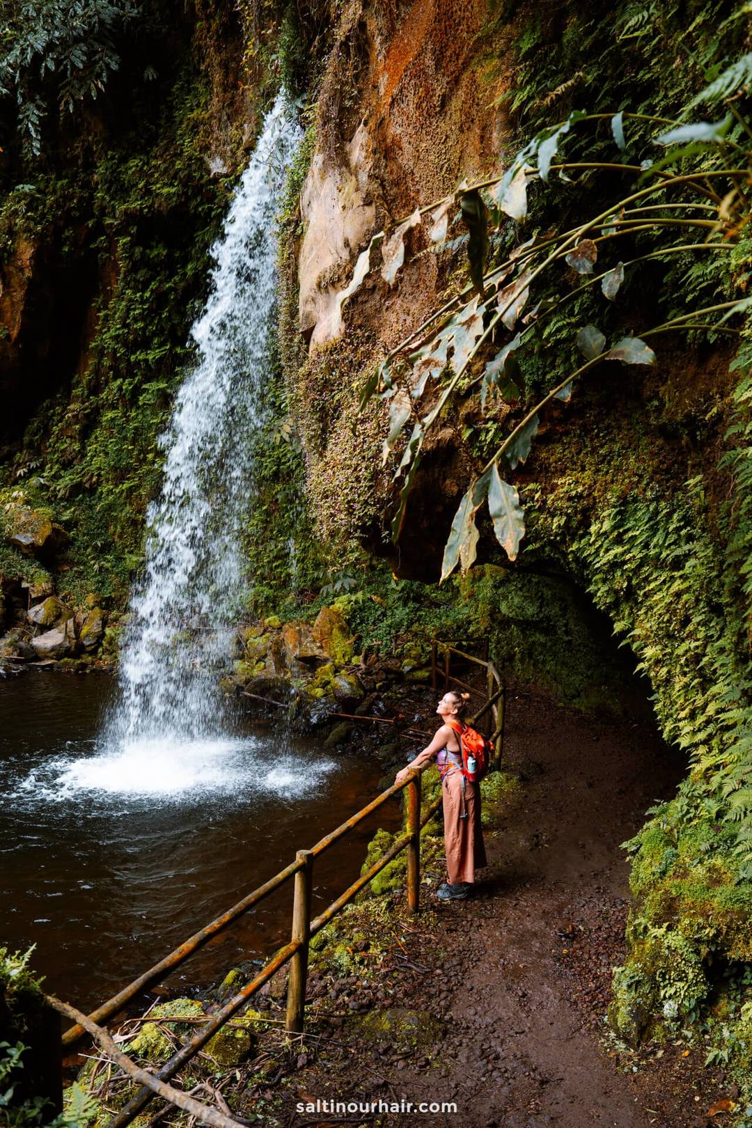 A woman with a red backpack stands on a muddy trail by a railing, looking at a waterfall cascading into a pool surrounded by lush greenery&mdash;Cascata da Gruta is one of the unforgettable things to do in S&atilde;o Miguel, Azores