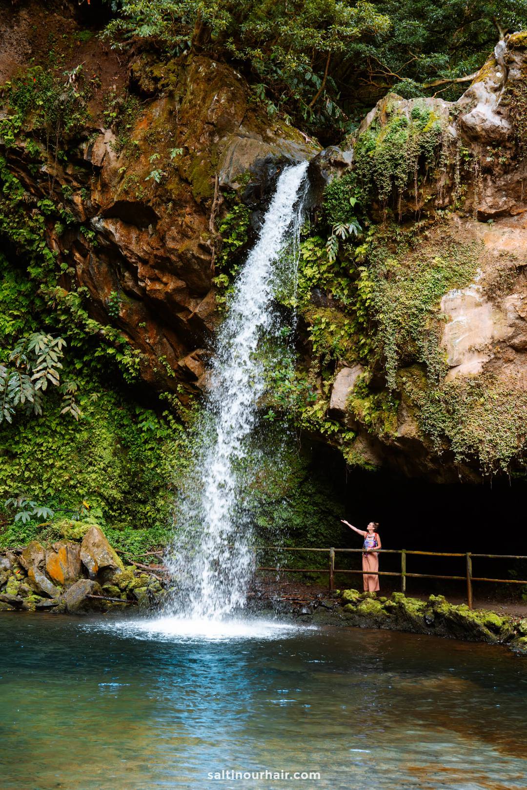 A woman stands on a railing next to a small waterfall called Cascata da Gruta, flowing from a rocky cliff surrounded by lush greenery into a clear pool&mdash;one of the enchanting things to do in S&atilde;o Miguel, Azores.