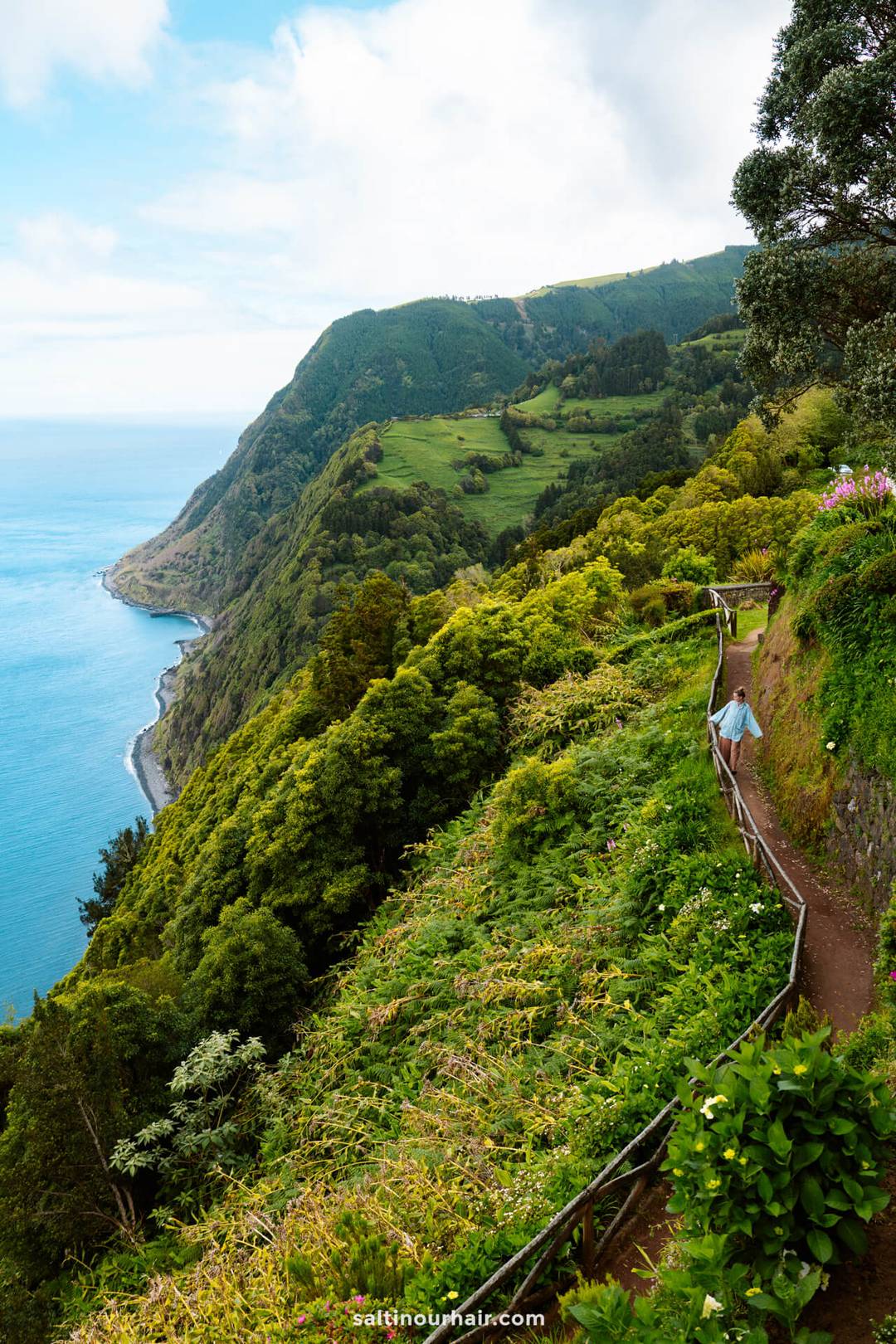 A woman walks along a narrow path surrounded by lush green cliffs overlooking the ocean, with a coastline and hills in the background at Miradouro da Ponta do Sossego&mdash;a perfect scene among the top things to do in S&atilde;o Miguel, Azores.