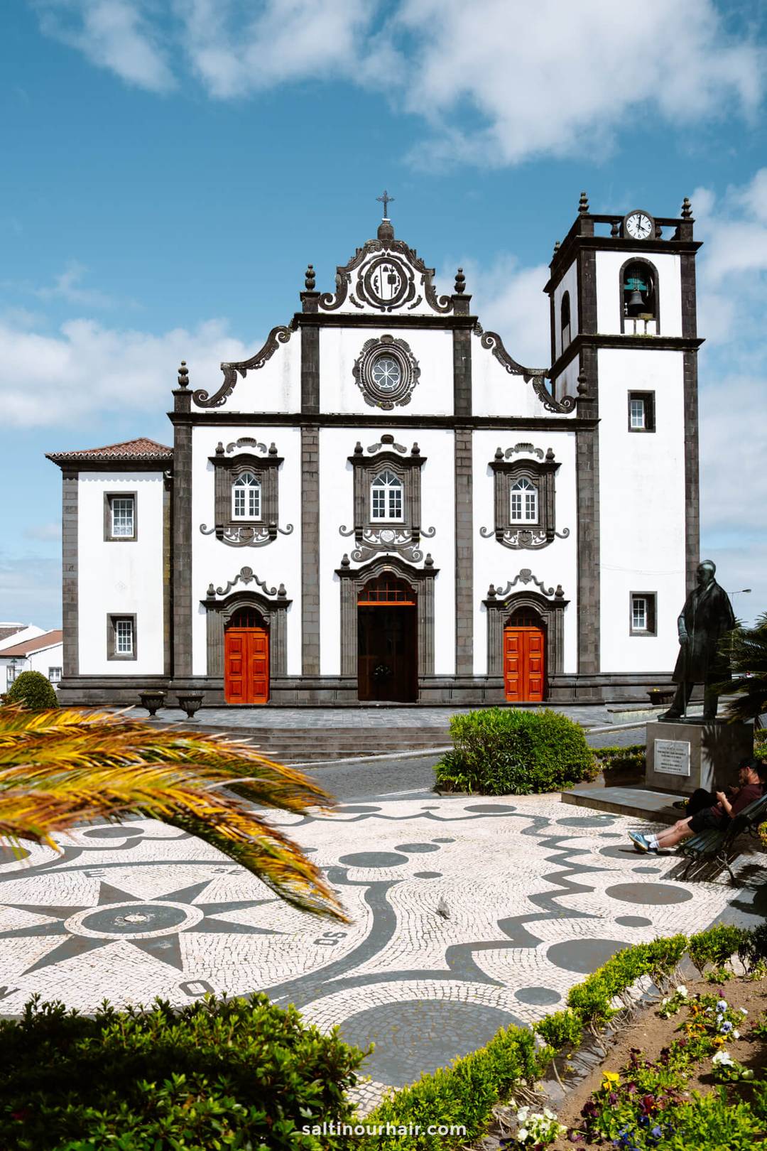 A white and black baroque-style church with red doors, a bell tower, and a patterned stone plaza in the foreground in Nordeste, S&atilde;o Miguel Azores.