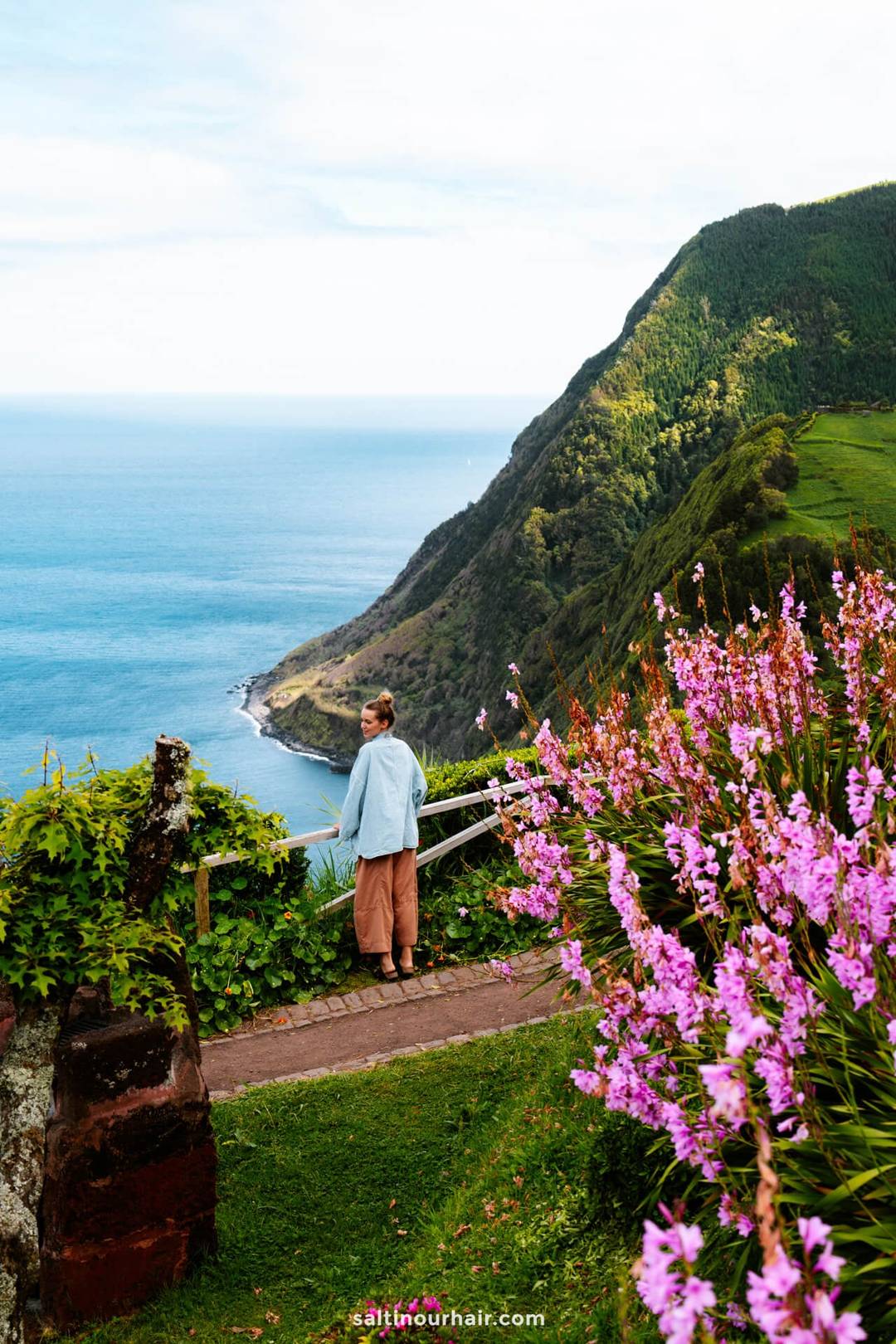 A woman stands on a path overlooking a coastal cliff at Miradouro da Ponta do Sossego, with pink flowers in the foreground and the ocean and green hills in the background&mdash;one of the breathtaking things to do in S&atilde;o Miguel, Azores.