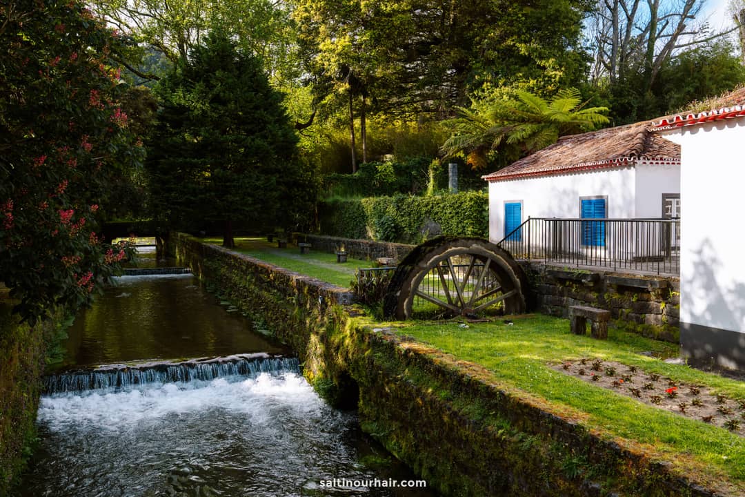 A small watermill with a wooden wheel sits beside a narrow stream surrounded by greenery and trees under daylight&mdash;a peaceful scene in the town of Furnas, S&atilde;o Miguel Azores.