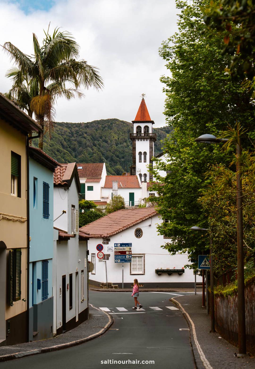 A woman crossed a narrow street with colorful houses in the small town of Frunas, with a church steeple and green hills visible in the background&mdash;capturing the charm of everyday life and inspiring ideas for things to do in S&atilde;o Miguel, Azores.