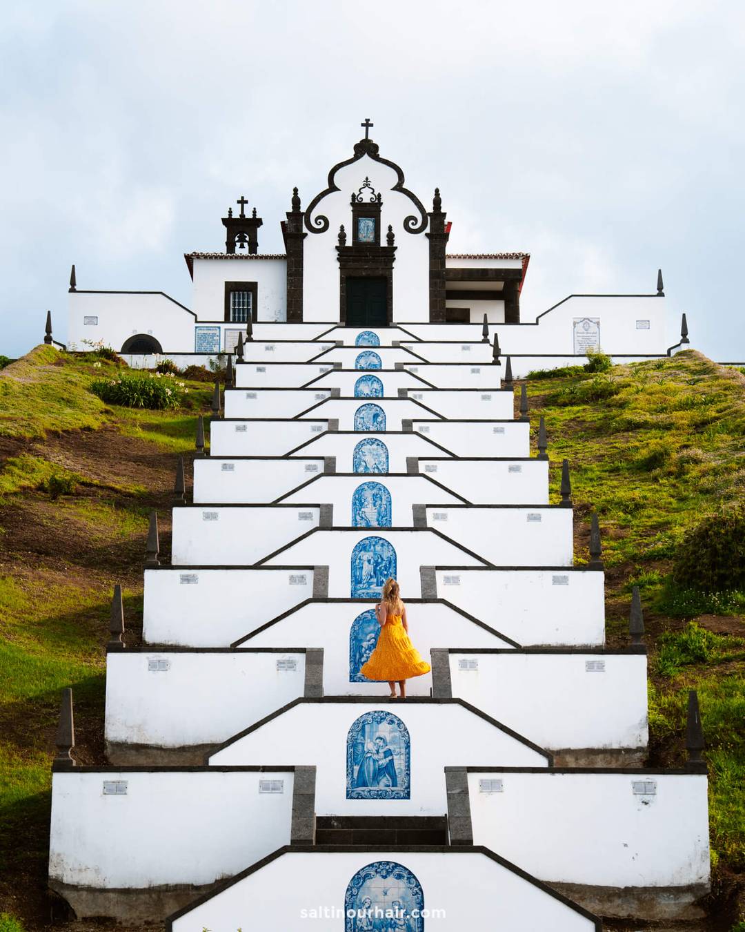 A woman in a yellow dress stands on the steps of a white church with blue tile artwork called Nossa Senhora da Paz Chapel&nbsp;in the town of Vila Franca&mdash;one of the picturesque things to do in S&atilde;o Miguel, Azores.