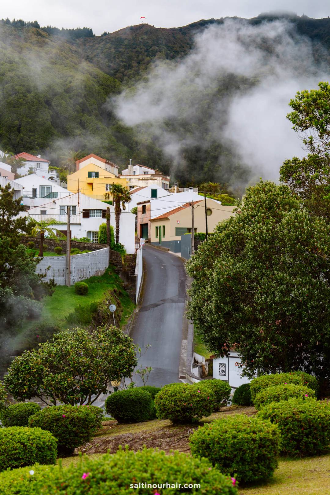 A narrow road leads through a lush, green village of Furnas with white and yellow houses, surrounded by volcanic steam and hills&mdash;an inviting scene among the many things to do in S&atilde;o Miguel, Azores.