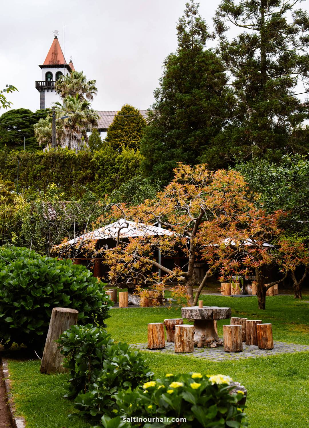 A garden restaurant with green grass, wooden tables and stools, leafy trees, and a church tower in the background&mdash;perfect for relaxing and discovering unique things to do in S&atilde;o Miguel, Azores.