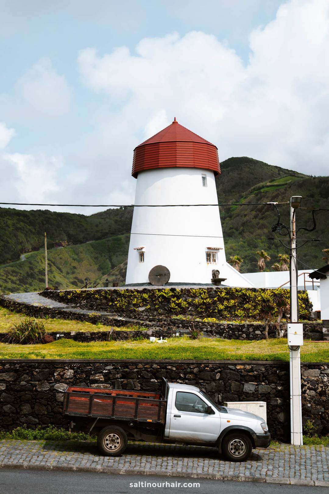 A white windmill with a red roof stands on a grassy hill beside a stone wall and parked pickup truck, set against mountains and a cloudy sky in S&atilde;o Miguel, Azores
